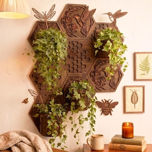Puede incluir: Un jardinera de pared de madera con diseño de panal, con colibríes, abejas y mariposas talladas. Las jardineras hexagonales contienen plantas verdes colgantes. También se ven grabados botánicos enmarcados y una vela encendida.