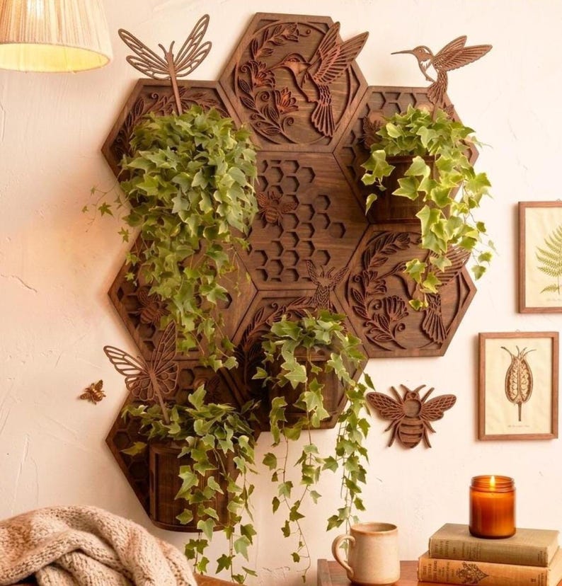 May include: A wooden wall planter with a honeycomb design, featuring hexagon-shaped shelves holding potted plants with trailing green vines. Decorative laser-cut hummingbirds, butterflies, and bees adorn the dark brown wood, creating a nature-inspired display.