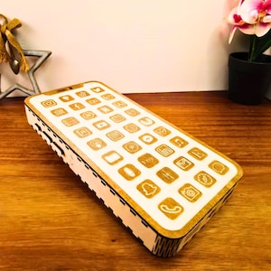 Könnte beinhalten: Eine Holzbox in Form eines Smartphones mit einer cremefarbenen Oberseite mit eingravierten App-Symbolen. Die Seiten sind helles Holz. Die Box steht auf einer Holzoberfläche.
