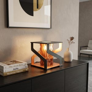 Puede incluir: Una lámpara de mesa de madera moderna con marco negro y luz cálida y brillante. La lámpara presenta un diseño único con elementos de madera en capas y se encuentra sobre una superficie de madera oscura. Hay libros apilados cerca.