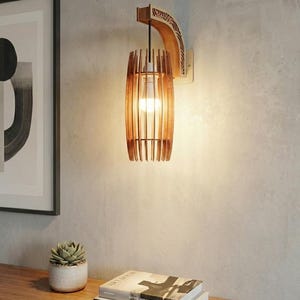 May include: A wooden wall sconce with a cylindrical shade made of vertical wooden slats. The sconce has a decorative wooden arm and a warm light bulb inside. The fixture is mounted on a light-colored wall, with a framed art print and a small succulent plant nearby.