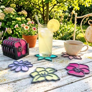 May include: A set of flower-shaped coasters in various colors including magenta, light purple, and pale yellow, stored in a black holder. The coasters feature a layered design with a dark outline and a solid center, displayed on a white wooden table.