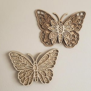 May include: Two wooden butterfly wall art pieces. The butterflies are layered with intricate cut-out designs, showcasing a natural wood grain finish. The butterflies are mounted on a neutral-colored wall, creating a decorative and artistic display.