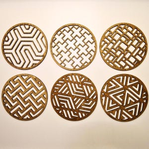 Puede incluir: Seis posavasos redondos de madera con intrincados diseños geométricos. Cada posavasos presenta un diseño único, con formas angulares y espacios abiertos. Los posavasos son de madera de color marrón claro.