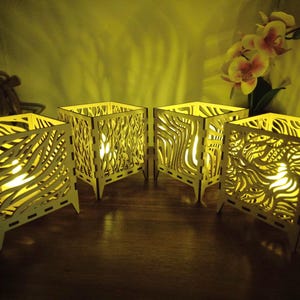 May include: Four square wooden candle holders with intricate cut-out designs. The candle holders are lit from within, casting a warm yellow glow. The designs vary, creating a unique decorative element. The candle holders are on a wooden surface.