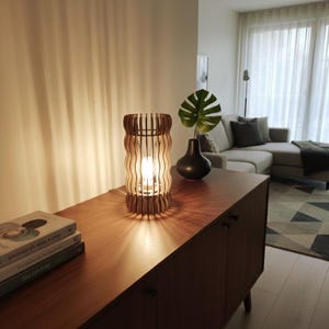 May include: A wooden table lamp with a wavy, vertical slat design. The lamp is lit, casting a warm glow. It sits on a dark wood cabinet, next to a black vase with a large green leaf. The room has a modern, minimalist aesthetic.