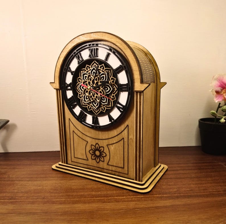 May include: Wooden mantel clock with a decorative floral design. The clock face features black Roman numerals on a white background, with a detailed black and gold mandala in the center. The clock's hands are red.