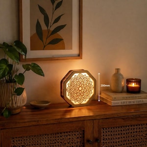 Puede incluir: Lámpara octogonal de madera con diseño de mandala, de 200 mm de altura. La lámpara está iluminada, emitiendo un brillo cálido. Se encuentra sobre un mueble de madera, junto a libros, una vela y un pequeño jarrón. Un grabado botánico enmarcado cuelga encima.