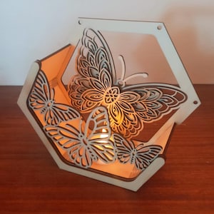 May include: A wooden, hexagonal candle holder with laser-cut butterfly designs. The butterflies are intricately detailed and arranged around the sides of the holder. The warm glow of a candle illuminates the interior, creating a soft, inviting ambiance.