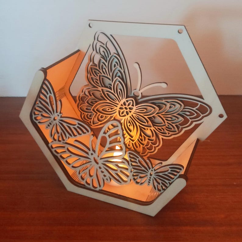 May include: A wooden, hexagonal candle holder with laser-cut butterfly designs. The butterflies are intricately detailed and arranged around the sides of the holder. The warm glow of a candle illuminates the interior, creating a soft, inviting ambiance.