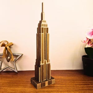 Puede incluir: Maqueta de madera del Empire State Building. El modelo está hecho de madera marrón clara y oscura, con líneas verticales y formas rectangulares. El modelo está sobre una superficie de madera. Un artículo decorativo.
