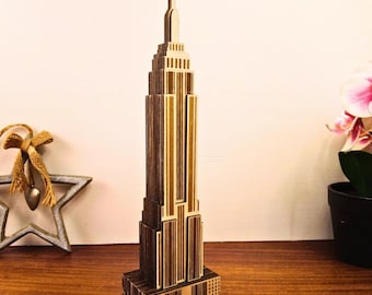 May include: A wooden Empire State Building model with a detailed design. The model is made of light and dark brown wood, with vertical lines and rectangular shapes. The model is standing on a wooden surface.