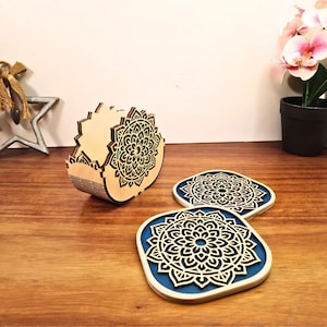May include: Wooden coasters with a laser-cut mandala design, presented with a matching holder. The coasters have a blue base. The holder and coasters are made of light-coloured wood. The coasters are approximately 10 cm in diameter.