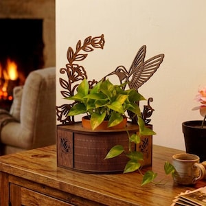 Peut inclure: Un cache-pot en bois brun foncé découpé au laser, orné d'une silhouette de colibri et de vignes fleuries. La base semi-circulaire présente un motif grillagé texturé et des découpes latérales. Un pot en terre cuite avec un pothos vert est inséré.