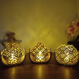 Op de afbeelding: Drie decoratieve houten kandelaars met ingewikkelde geometrische uitsparingen. Elke houder heeft een warm, gloeiend licht van binnen, waardoor een gezellige sfeer ontstaat. De houders zijn gerangschikt op een houten oppervlak, met een stervormig ornament en een orchidee in pot op de achtergrond.