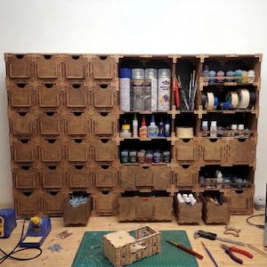 May include: A large wooden storage unit with many small drawers and open shelves. The unit is filled with various craft supplies, including spray paint cans, small bottles of paint, rolls of tape, and tools. The drawers have cut-out handles.