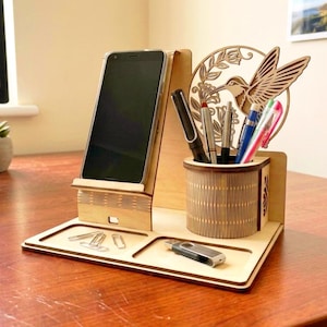 Puede incluir: Un organizador de escritorio de madera clara que incluye un soporte vertical para smartphone, un portalápices cilíndrico con un diseño de colibrí y flores cortado con láser, y dos bandejas empotradas con clips metálicos y una memoria USB plateada.