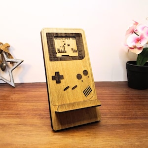 May include: Wooden phone stand shaped like a retro handheld game console. The stand features a screen displaying a pixelated game scene, a directional pad, and other buttons. The wood has a light brown color.