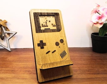 Könnte beinhalten: Hellbrauner Handy-Ständer aus Holz in Form einer Retro-Handheld-Spielkonsole. Der Bildschirm zeigt eine pixelige Spielszene. Er hat ein Steuerkreuz, Knöpfe und einen Lautsprecher.