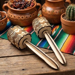 May include: Two wooden percussion instruments with cylindrical bodies featuring laser-etched geometric patterns. Each instrument has a long, flat handle extending from the centre of the cylinder, constructed from light-coloured wood with dark edges.