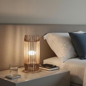 Puede incluir: Lámpara de mesa de madera con diseño cilíndrico, con listones verticales y una bombilla iluminada. La lámpara está sobre una mesita de noche blanca junto a un vaso de agua y un libro. La lámpara mide unos 30 cm de alto.