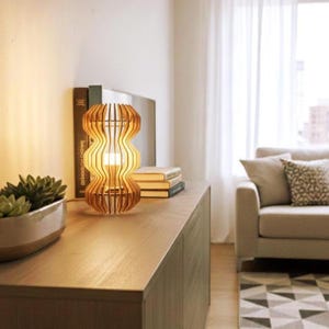 Może przedstawiać: Drewniana lampa stołowa o unikalnym, falistym designie. Lampa wykonana jest z jasnego drewna i ma ciepłą, zachęcającą poświatę. Lampa stoi na drewnianej szafce, w pobliżu znajdują się książki i roślina sukulent. W tle widoczna jest sofa i okno.