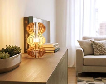 Könnte beinhalten: Eine Holz-Tischlampe mit einem einzigartigen, wellenförmigen Design. Die Lampe besteht aus hellem Holz und hat einen warmen, einladenden Schein. Sie steht auf einem Holzschrank, mit Büchern und einer Sukkulentenpflanze in der Nähe. Im Hintergrund sind ein Sofa und ein Fenster zu sehen.