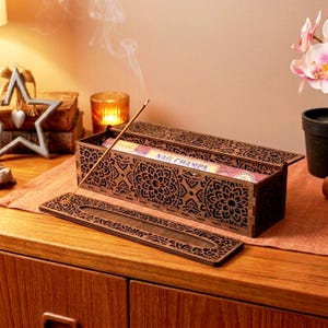 Puede incluir: Un porta incienso de madera con intrincados calados florales. La caja está abierta, revelando varillas de incienso etiquetadas como "Nag Champa". Una varilla de incienso encendida emite humo. El soporte está sobre una superficie de madera.