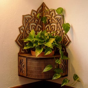 Op de afbeelding: Een donkerbruine houten hoekplank met een ingewikkeld lasergesneden mandalapatroon op het achterpaneel en een lattenontwerp aan de voorzijde. Een groene pothosplant in een terracotta pot staat op de plank, met naar beneden hangende ranken.