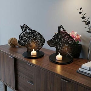 May include: Two black metal candle holders shaped like a cat and a wolf, each with intricate floral designs. The candle holders sit on black circular bases, each holding a lit white tea light candle. The items are displayed on a wooden surface.