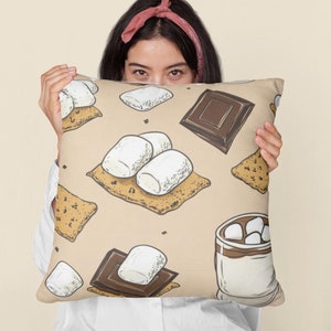 May include: A beige pillow with a pattern of chocolate squares, graham crackers, and marshmallows. The pillow is being held by a person with a pink headband.