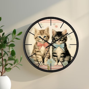 Puede incluir: Un reloj de pared redondo con marco negro y fondo blanco. La esfera del reloj presenta una ilustración vintage de dos gatitos con pajaritas. Los gatitos están sentados uno al lado del otro y mirando al espectador.
