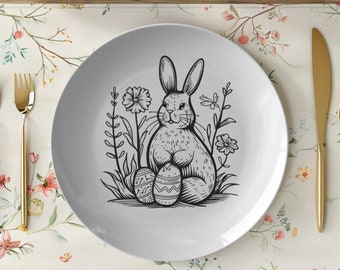 May include: A white ceramic plate with a black and white illustration of a rabbit holding Easter eggs. The plate is on a floral placemat with gold silverware.
