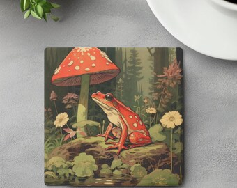 May include: A square coaster featuring a vibrant illustration of a red frog with white spots sitting on a rock. A large red mushroom with white spots stands behind the frog in a forest setting with green foliage, small yellow flowers, and water.