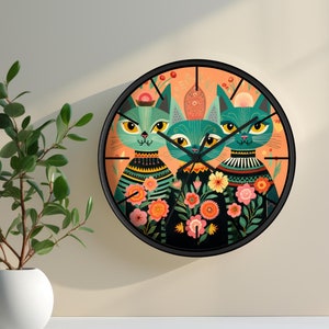 Könnte beinhalten: Eine runde Wanduhr mit schwarzem Rahmen und einem farbenfrohen Design mit drei stilisierten Katzen mit floralen Akzenten. Der Hintergrund ist in einem Pfirsich-Orange gehalten.