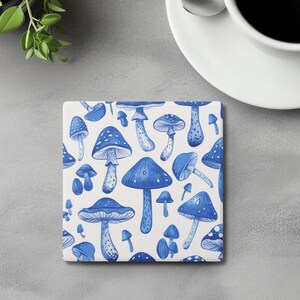 May include: Square ceramic coaster featuring a pattern of blue mushrooms on a white background. The mushrooms vary in size and shape, with detailed caps and stems. A white coffee cup with black coffee sits on a saucer next to the coaster.