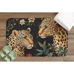 May include: A rectangular bath mat featuring a black background with two leopards, white flowers, and green foliage. The mat is positioned on a wooden floor next to a pair of light green sandals and a folded white towel.