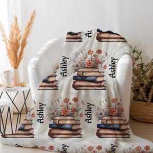 May include: A cream-colored throw blanket featuring a repeating pattern of stacked books and floral arrangements. The name "Ashley" is printed vertically on the blanket. The blanket is draped over a white armchair.