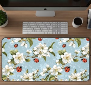 May include: Rectangular desk mat featuring a floral design with white blossoms, green leaves, and red ladybugs on a light blue background. The mat is designed to protect the desk surface and provide a smooth surface for a mouse and keyboard.