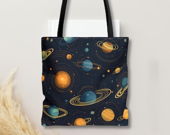 Op de afbeelding: Een donkerblauwe tote bag met een herhalend patroon van diverse planeten, waaronder geringde gasreuzen en kleinere bollen in tinten oranje, geel en blauw, geaccentueerd door dunne gouden orbitale lijnen en kleine witte sterpuntjes.