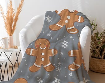 Puede incluir: Una manta gris suave con un patrón repetitivo de hombres de jengibre marrones con detalles de glaseado blanco y pequeños copos de nieve blancos esparcidos por la tela. Las figuras de jengibre tienen caras sonrientes y botones blancos.