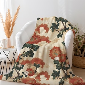 Könnte beinhalten: Eine cremefarbene Decke mit einem Blumenmuster in Rot-, Orange- und Grüntönen. Die Decke ist über einen weißen Stuhl drapiert. Das florale Design umfasst große Blumen und belaubte Zweige und schafft eine Vintage-Ästhetik.