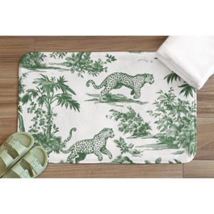May include: A rectangular bath mat featuring a white background with a repeating green toile pattern. The design displays two leopards in motion surrounded by detailed illustrations of tropical palm trees, lush foliage, and various leafy plants.