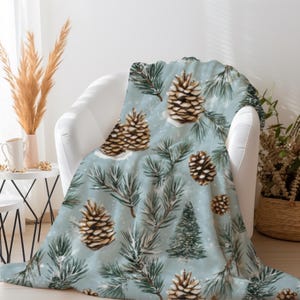 May include: A soft throw blanket featuring a light blue background with a repeating pattern of brown pinecones, green pine branches, and small evergreen trees. The design includes white speckles that resemble falling snow across the fabric.