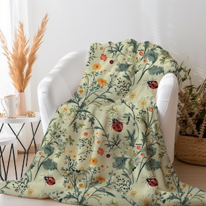 May include: A soft, green throw blanket with a floral pattern featuring red ladybirds and yellow flowers. The blanket is draped over a white armchair.
