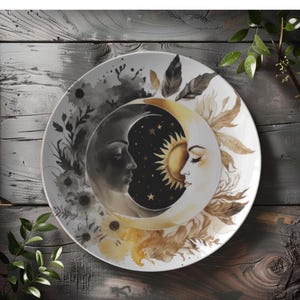 May include: A white ceramic plate featuring a celestial design. The plate is divided into two halves, one depicting a crescent moon and the other a sun, both with human faces. The design includes stars, floral elements, and a color palette of black, gold, and white.