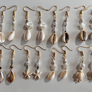 May include: A set of 10 pairs of gold earrings featuring a variety of charms, including seashells, stars, pearls, and a turtle. The earrings are all made with gold-toned metal and have a delicate, feminine design.