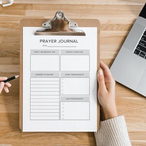 May include: A white prayer journal with a brown clipboard. The journal has sections for gratitude, prayer requests, and answers to prayers. The journal is on a wooden table with a laptop and a pair of headphones.