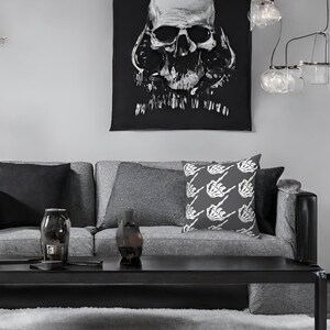 May include: A gray couch with a black skull tapestry hanging above it. The couch has two gray throw pillows and one throw pillow with a white skeleton hand pattern. The couch is in front of a gray wall.