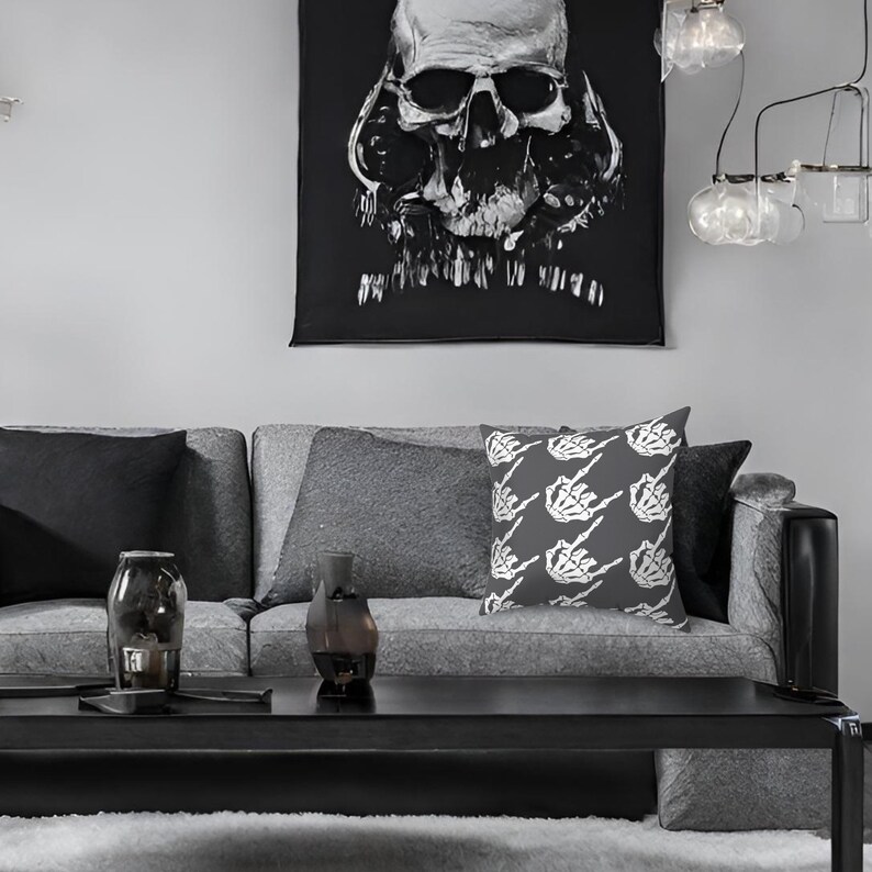 May include: A gray couch with a black skull tapestry hanging above it. The couch has two gray throw pillows and one throw pillow with a white skeleton hand pattern. The couch is in front of a gray wall.